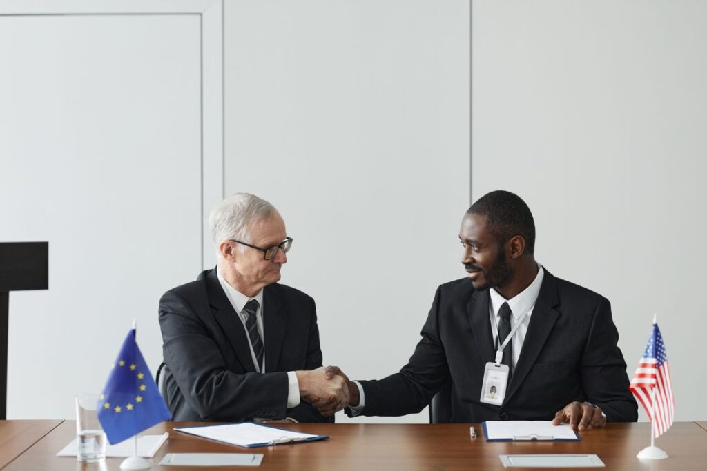 Zwei Diplomaten schuetteln sich am Verhandlungstisch mit EU- und US-Flagge die Haende
